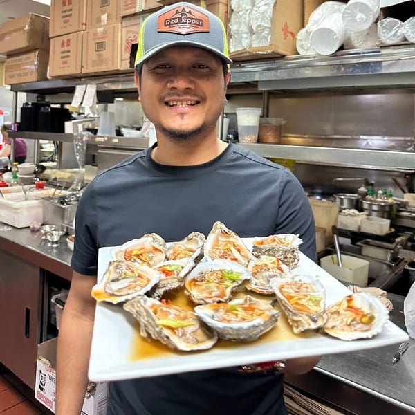 A smiling chef in a busy Asian Bistro kitchen holds a platter of fresh oysters with sauce.