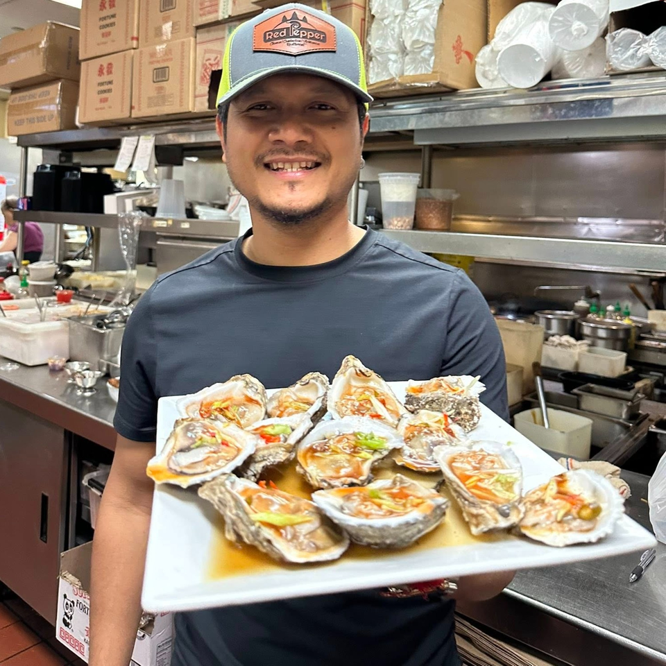 A smiling chef in a busy Asian Bistro kitchen holds a platter of fresh oysters with sauce.