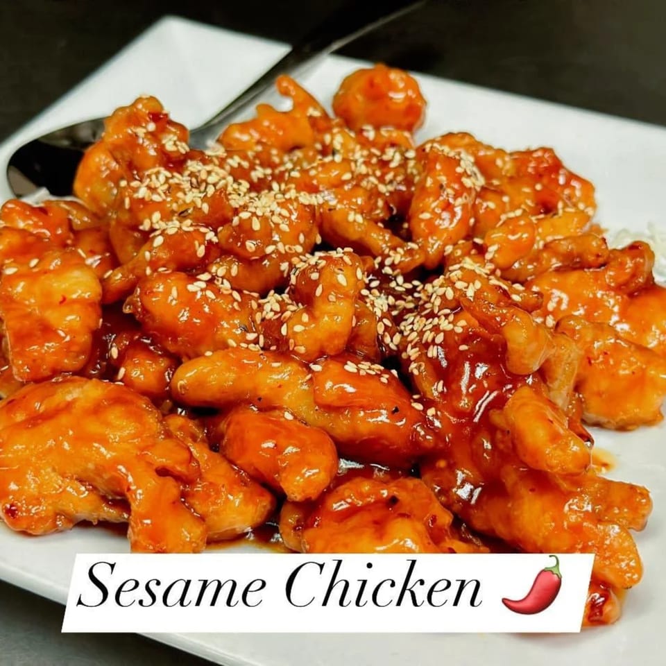 Sesame chicken, coated in a glossy orange sauce and sprinkled with sesame seeds, on a white plate.