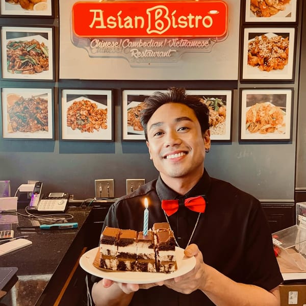 Smiling man in a red bow tie holding a plate of chocolate cake slices with a lit candle.